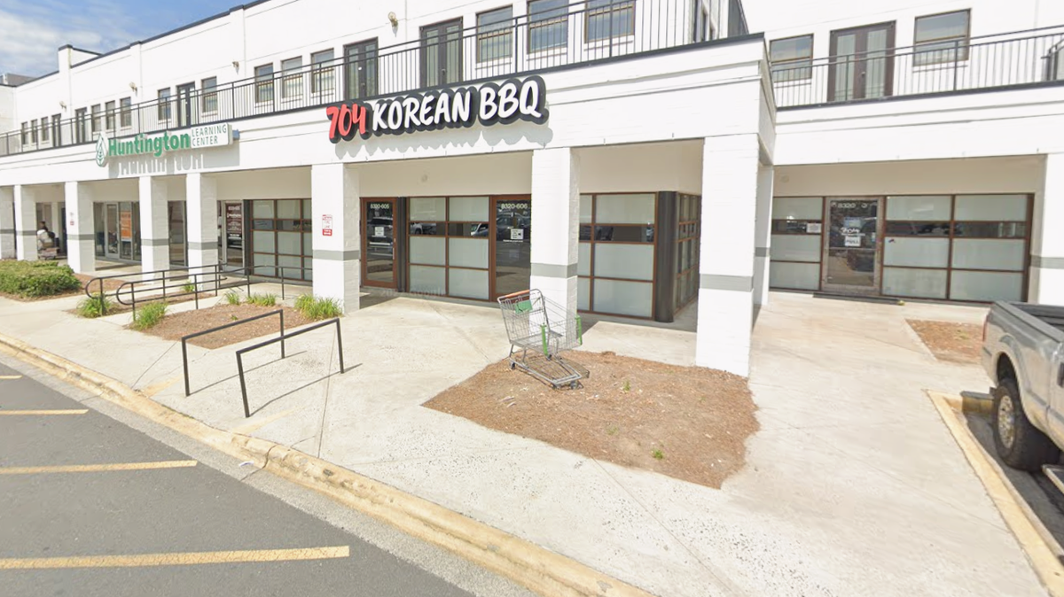 The exterior of the 704 Korean BBQ restaurant, which has a red and white sign. The restaurant is located in a white brick strip mall next to a Huntington Learning Center. The image also shows a sidewalk, some planted dirt patches, and a metal shopping cart resting in front of the main entrance.
