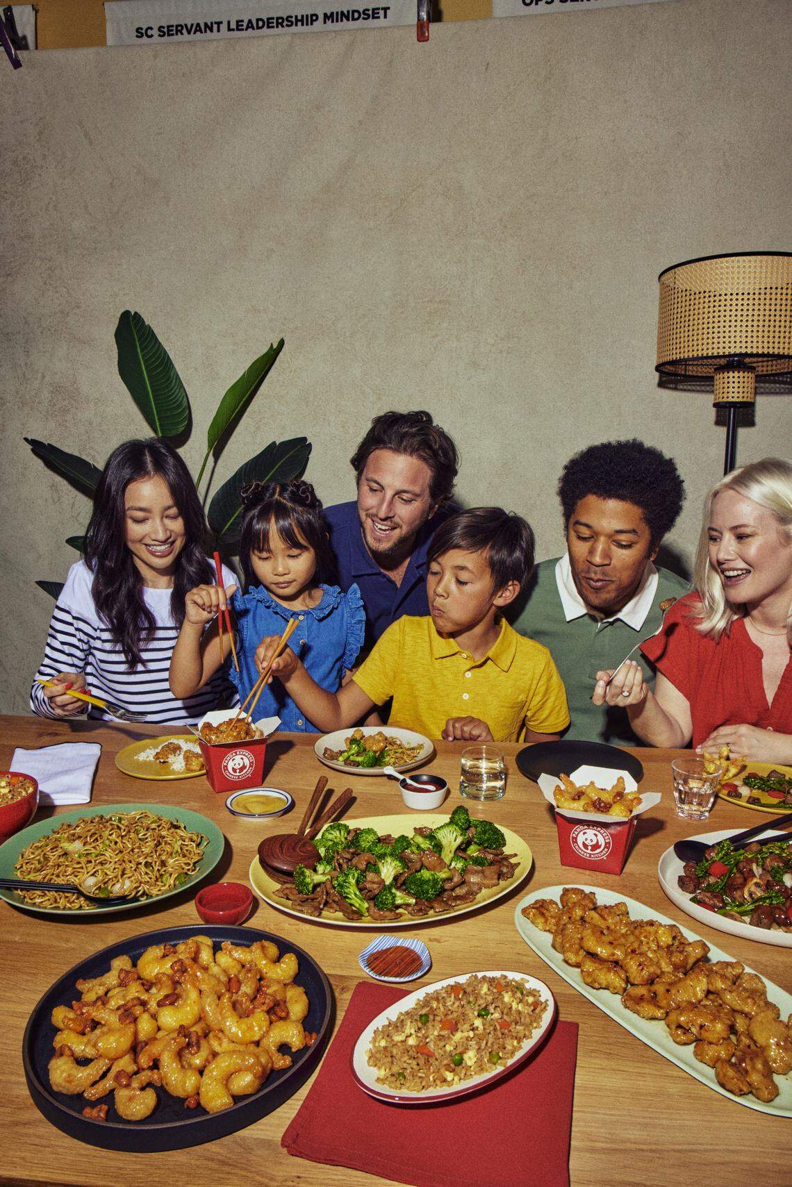A diverse group of family and friends gather around a wooden table to share a large meal of Chinese-American favorites. The scene captures a joyful moment of connection as children and adults use chopsticks and forks to serve themselves from various platters.