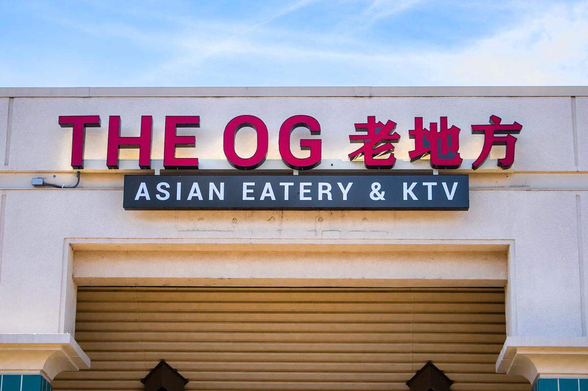 An exterior shot of a storefront sign for “THE OG 老地方” in large red letters, with the subtitle “ASIAN EATERY & KTV” in white letters on a black background below it, against a cream-colored building.