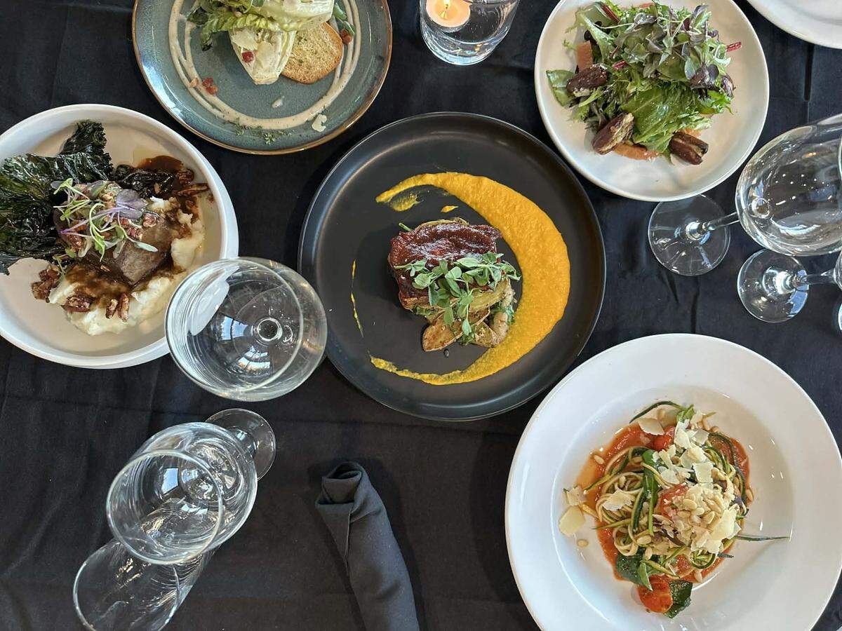 A top-down view of a full dinner spread on a black tablecloth, showcasing five gourmet plates such as pasta with pine nuts and a salad with dates. The arrangement includes multiple wine glasses and a black linen napkin, highlighting a diverse and sophisticated multi-course meal.