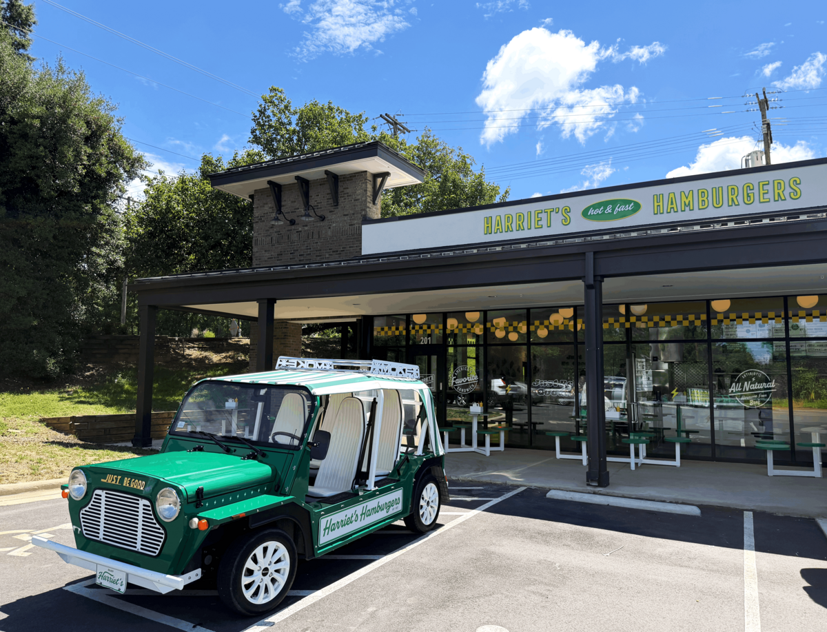 The exterior of Harriet’s Hamburgers is shown on a bright, sunny day under a blue sky with fluffy white clouds. The modern building features a dark awning, large glass windows with green outdoor seating underneath, and a sign reading “Harriet’s Hot & Fast Hamburgers.” A custom green and white vintage-style golf cart with the restaurant’s branding is parked in the foreground.