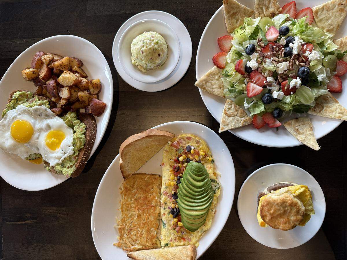 An overhead view of a dark wooden table covered with four white plates of various breakfast and lunch items, including avocado toast, an omelet with avocado slices, a fruit and nut salad with pita wedges, a side of potato salad, and a breakfast sandwich on a biscuit.