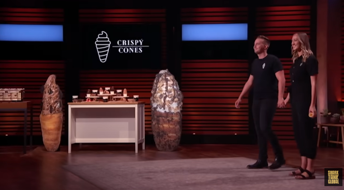 Two entrepreneurs, a man and a woman, stand smiling on a stage after walking toward the center. Behind them is a large screen displaying the “Crispy Cones” logo and a display table with samples of their product, flanked by two large, cone-shaped props.