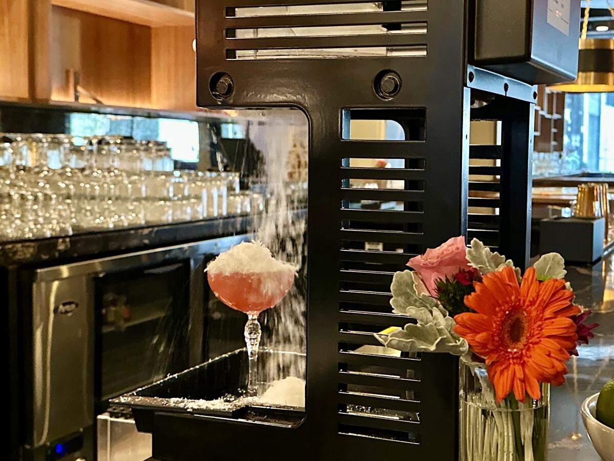 A black industrial ice shaver sits on a bar counter, mid-operation as it showers a pink cocktail with fine, white shaved ice. The elegant coupe glass is nearly topped with a snowy mound, while a vibrant bouquet of orange and pink flowers sits in the foreground. In the blurred background, rows of clean glassware and warm wooden cabinetry create a sophisticated bar atmosphere.