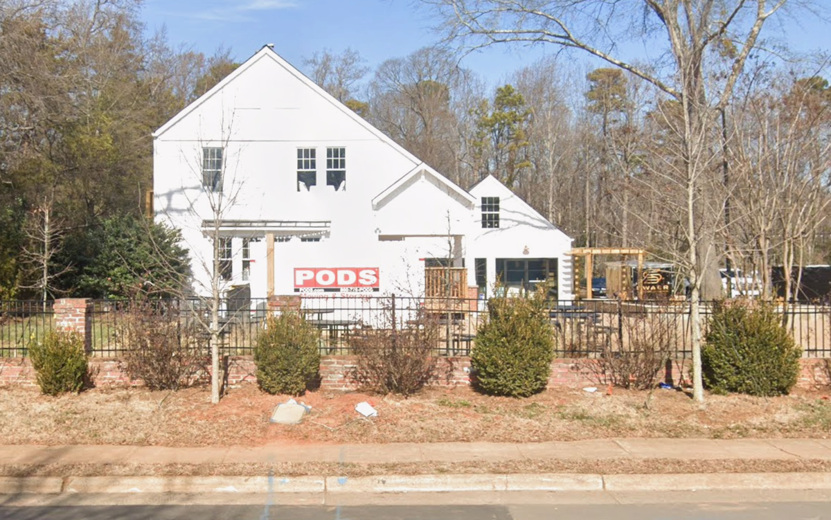 A two-story white building with a steeply pitched roof is shown behind a black wrought-iron fence and several small evergreen shrubs. A PODS moving and storage container is parked directly in front of the building, which is surrounded by leafless trees under a clear blue sky.