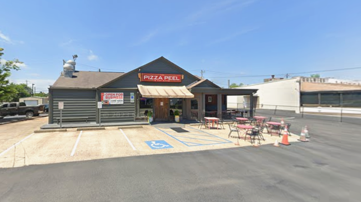 A small, single-story restaurant called The Pizza Peel & Tap Room is shown on a sunny day. The gray building has a brown awning and a small patio area with pink tables and red chairs in front, adjacent to a few parking spaces.