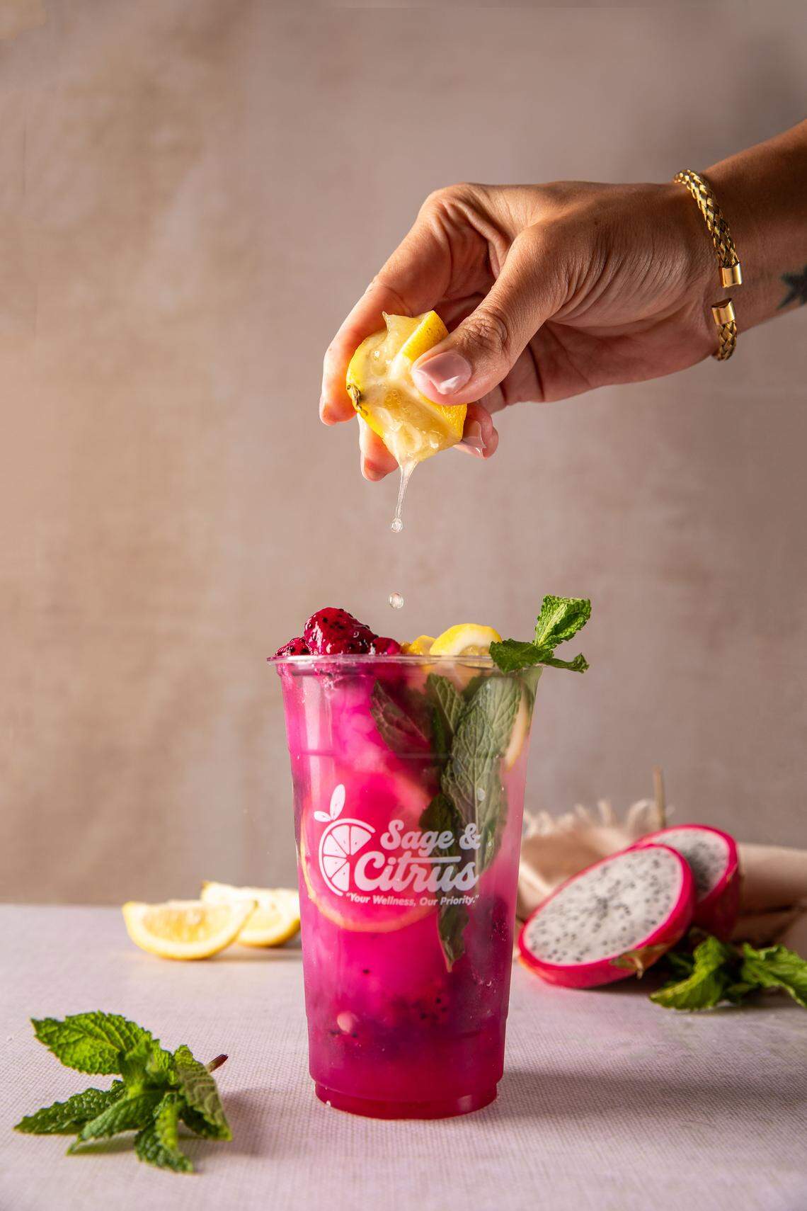 A hand squeezes a lemon wedge into a bright pink drink filled with mint leaves and chunks of dragon fruit. The refreshing beverage, labeled “Sage & Citrus,” is surrounded by mint and dragon fruit slices on a light-colored table.