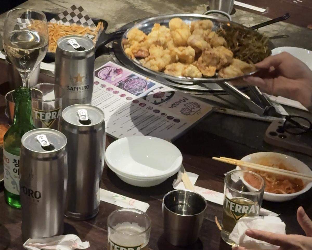 A crowded dinner table is filled with tall silver cans of Sapporo beer, glasses of Terra beer, and a bottle of Soju. In the center, a large platter of golden-brown fried chicken sits alongside small bowls of kimchi and noodles.