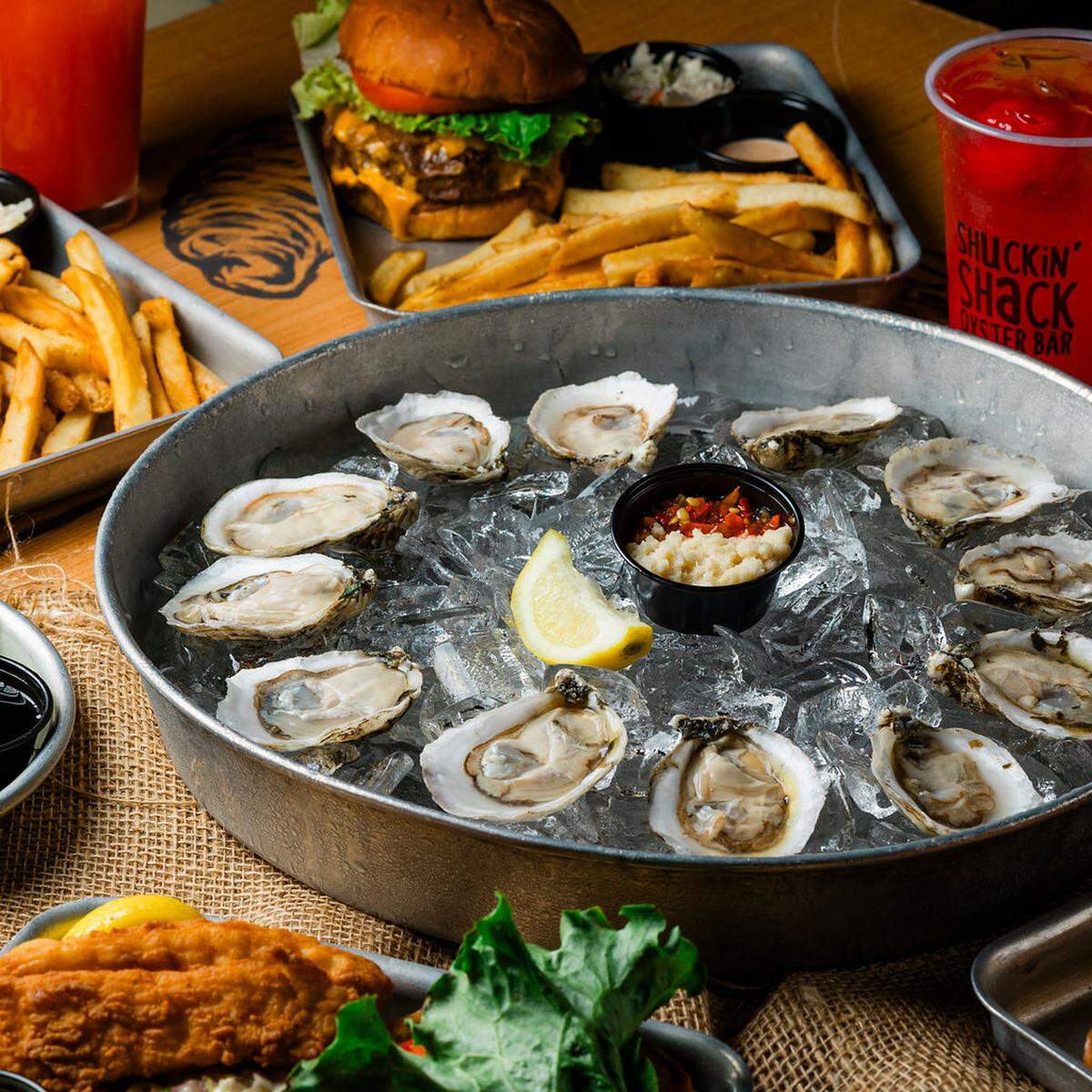 A close-up shot focuses on a metal basin of raw oysters served on a bed of crushed ice with a center tin of cocktail sauce and horseradish. In the blurred background, a hearty cheeseburger with fries and a red cocktail sit on a wooden table.