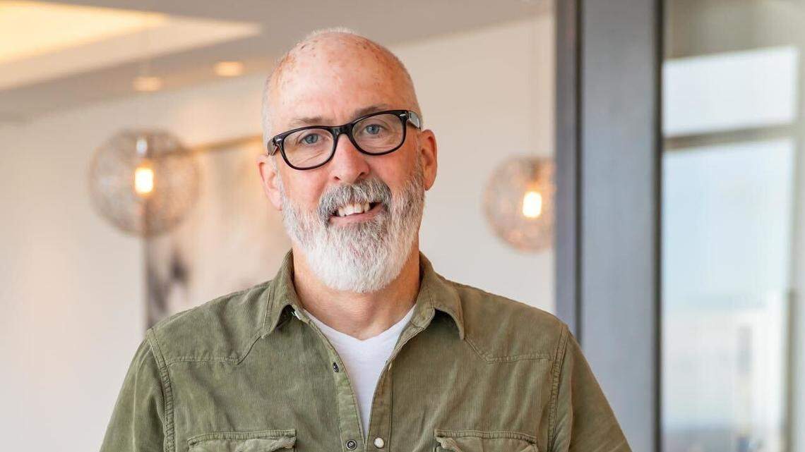 A smiling man with a grey beard and glasses, wearing a green corduroy shirt and blue jeans, stands leaning on a white desk in a modern office with large windows and light fixtures in the background.