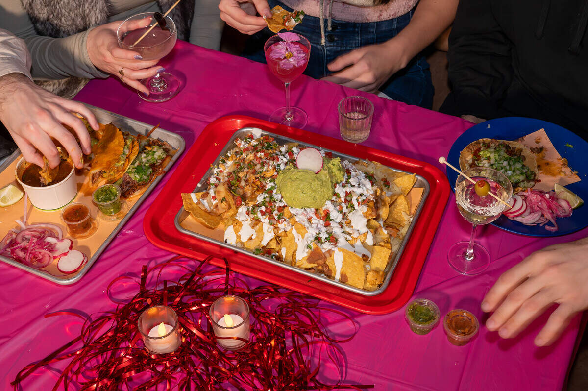 A vibrant overhead view captures a table covered in a bright pink tablecloth, laden with trays of loaded nachos, tacos, and colorful cocktails. Friends are seen sharing the meal, surrounded by festive red tinsel and small flickering candles.
