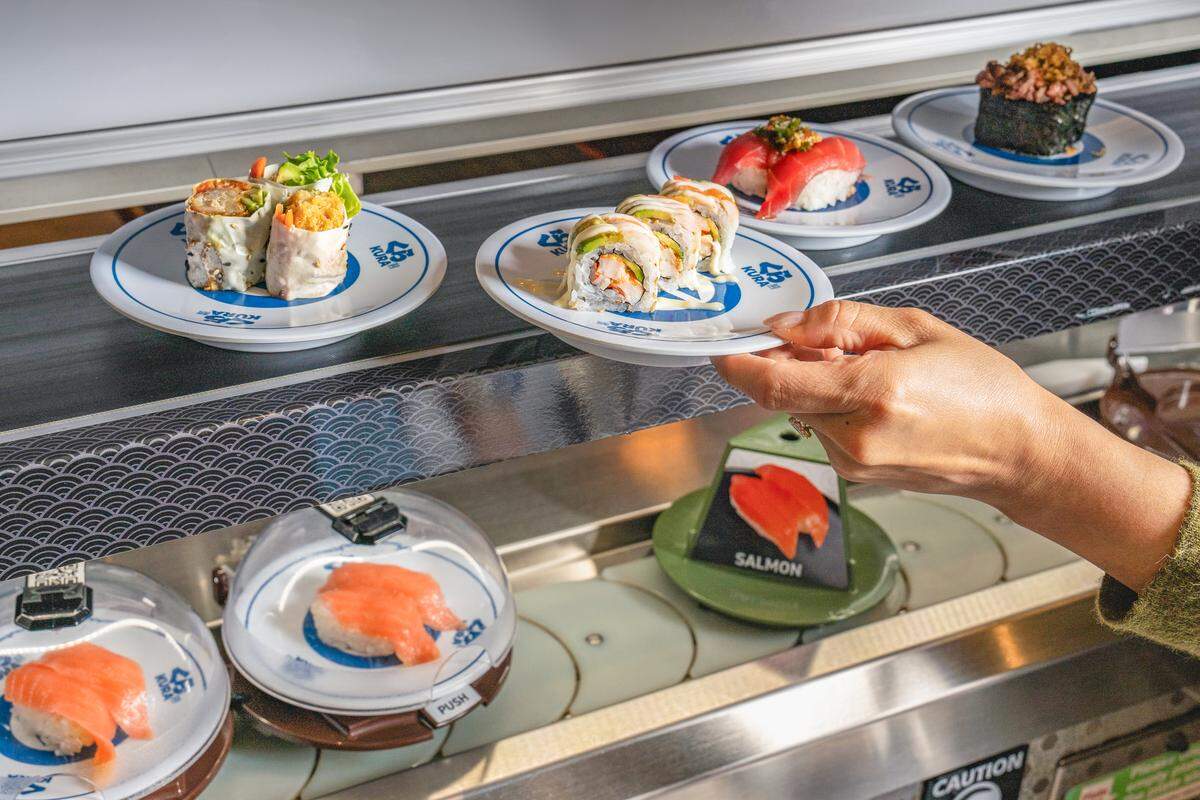 A hand reaches out to select a plate of avocado-topped sushi rolls from a moving double-decker conveyor belt system. The top tier features various rolls and nigiri on open plates, while the lower belt shows salmon nigiri protected by clear plastic domes. The industrial-style revolving bar is labeled with small signs identifying the different types of fish.