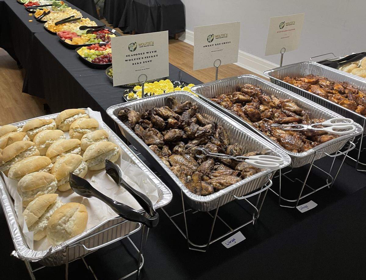 A catering spread is presented across two long tables, with the foreground featuring trays of BBQ wings, possibly a sauced meat dish, and a tray of small white sandwiches. The background table holds a variety of colorful fruit and cheese platters, with small signs displaying food labels for the event.