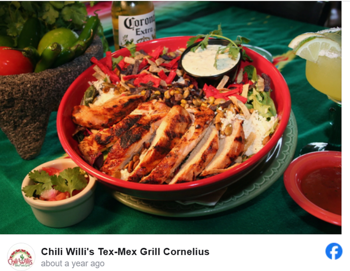 A large red bowl filled with a colorful Southwest grilled chicken salad sits at the center of a festive table spread. The salad features sliced charred chicken breast over a bed of greens, topped with roasted corn, black beans, pepitas, and vibrant red tortilla strips. The surrounding scene includes a Corona beer, a margarita, fresh salsa, and a molcajete filled with whole peppers and tomatoes on a green tablecloth.