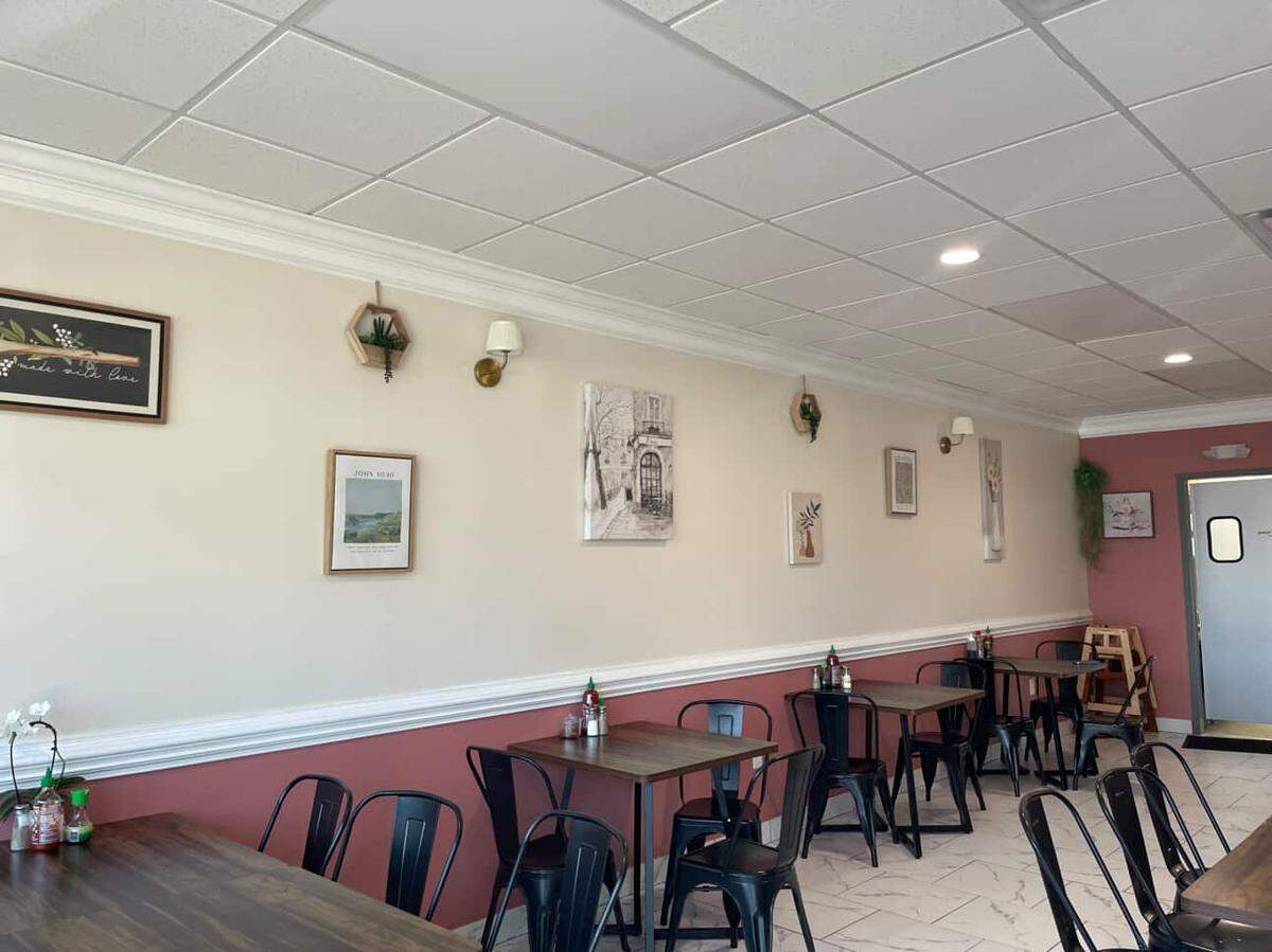 The restaurant interior features a warm, inviting atmosphere with wooden tables, black metal chairs, and walls decorated with framed art and small plants. Large windows and overhead lighting brighten the space, which includes a mix of white and rose-colored wall panels.