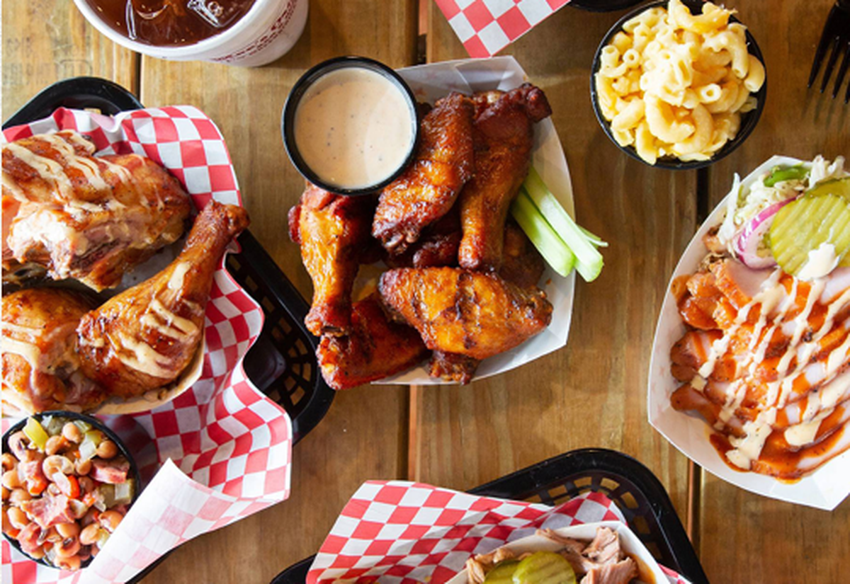 A top-down view shows a wooden table overflowing with barbecue favorites, including baskets of smoked wings, drumsticks drizzled with white sauce, and pulled pork. The spread is accompanied by classic Southern sides like macaroni and cheese, black-eyed peas, and fresh coleslaw with pickles. Red and white checkered paper liners add a vibrant, casual feel to this communal feast.