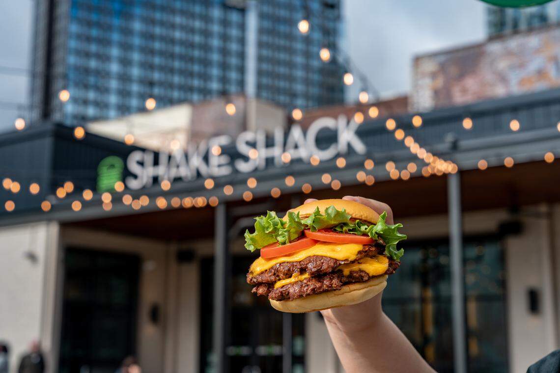 A person holds up a double ShackBurger with two beef patties, melted cheese, lettuce, and tomato against a blurred urban background. In the distance, a modern Shake Shack storefront is visible with its signature green logo and decorative string lights.