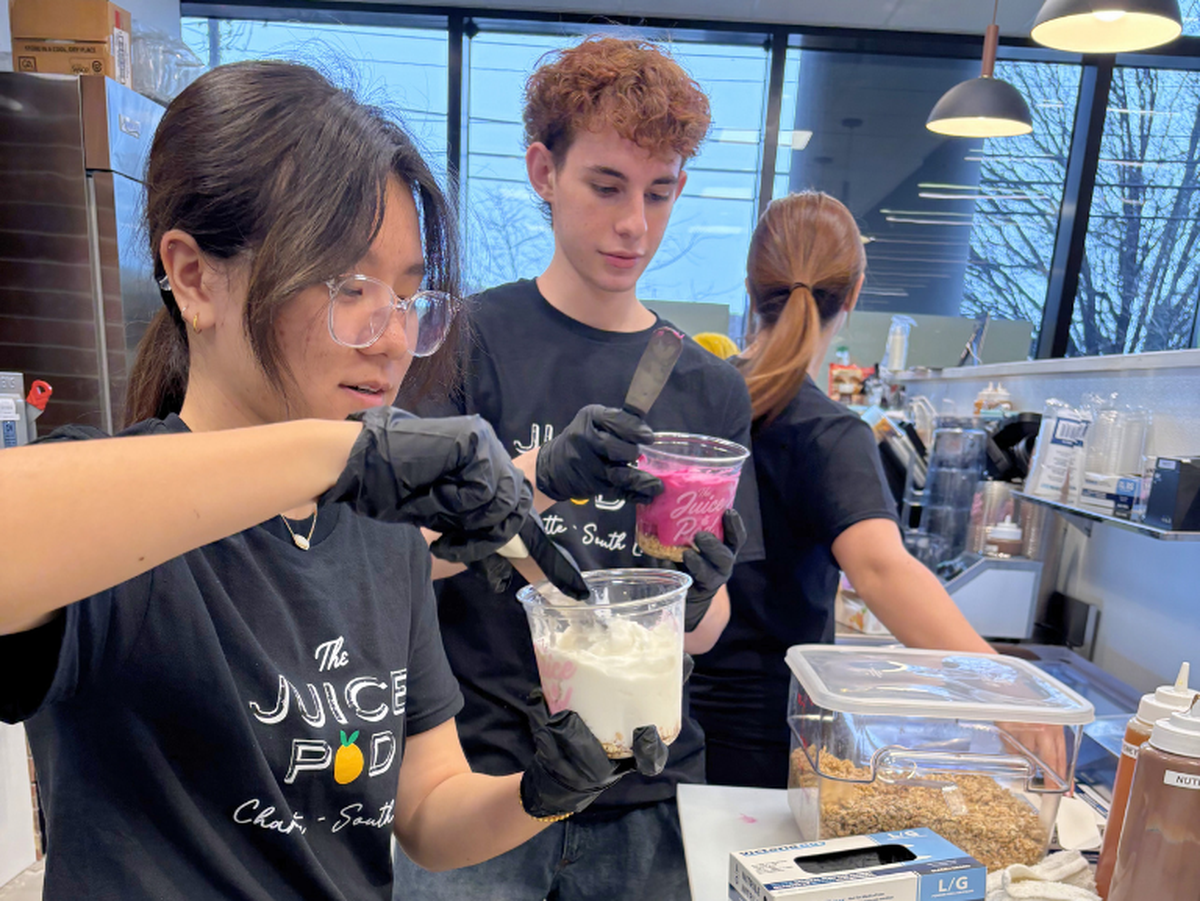 Two employees in black branded T-shirts and gloves carefully prepare smoothie bowls behind a cafe counter during a grand opening event. They are focused on layering fresh ingredients into clear plastic cups in a bright, modern kitchen setting.