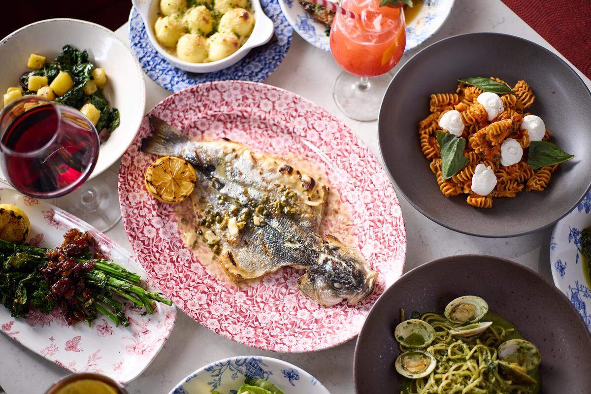 This vibrant bird’s-eye view captures a full dinner table laden with various Italian specialties including pasta, grilled fish, and colorful cocktails. Centered on a red-and-white floral platter is a large, herb-crusted whole fish accompanied by bowls of pesto linguine with clams and fusilli with tomato and mozzarella. The composition is lively and warm, emphasizing a shared dining experience with a variety of textures and colors.