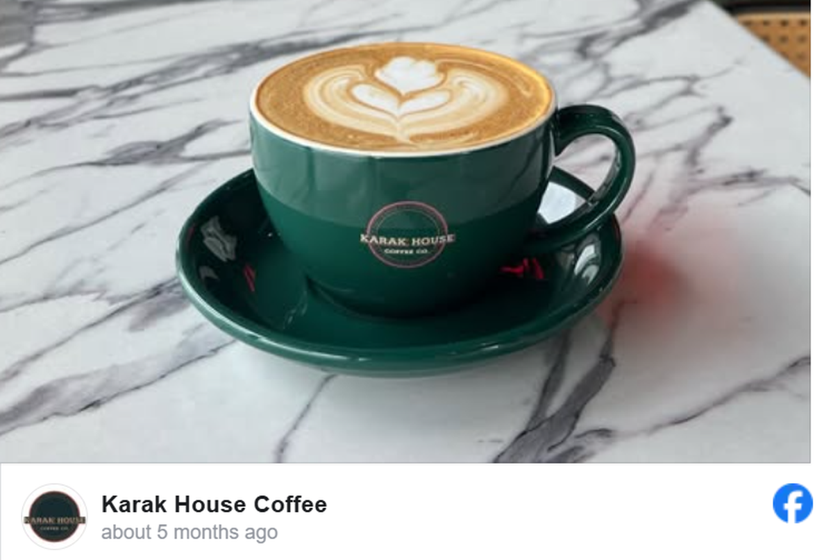 A steaming latte with intricate floral latte art is served in a dark green ceramic cup and saucer branded with the Karak House Coffee Co. logo. The cup sits atop a polished white marble tabletop with grey veining, creating a clean and sophisticated aesthetic. This image is a social media post from Karak House Coffee, appearing alongside their profile icon and a Facebook logo.