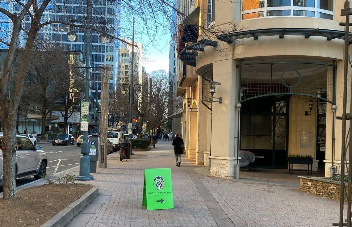 A bright green sandwich board with the Gotcha Matcha panda logo sits on a brick sidewalk to guide pedestrians toward the shop. The background shows a sunny city street in Charlotte with tall buildings, bare winter trees, and a person walking in the distance.