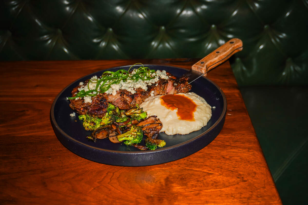 A seared 12oz Angus ribeye steak topped with blue cheese crumbles and scallions sits on a dark plate alongside mashed potatoes with gravy and roasted broccoli and mushrooms. The dish is presented on a wooden table in front of a dark green, tufted leather banquette.