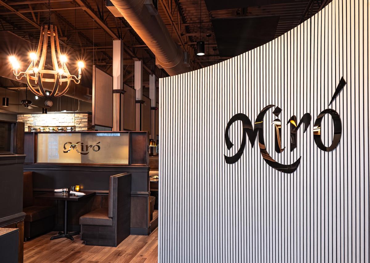 The restaurant entrance features a striking white wall with vertical black slats and a large, black script logo that reads “Miró.” The view looks past the entryway into the dining area, highlighting the warm wood flooring, dark booths, and industrial ceiling. A large wooden chandelier hangs in the distance, adding a rustic touch to the sophisticated, contemporary design.