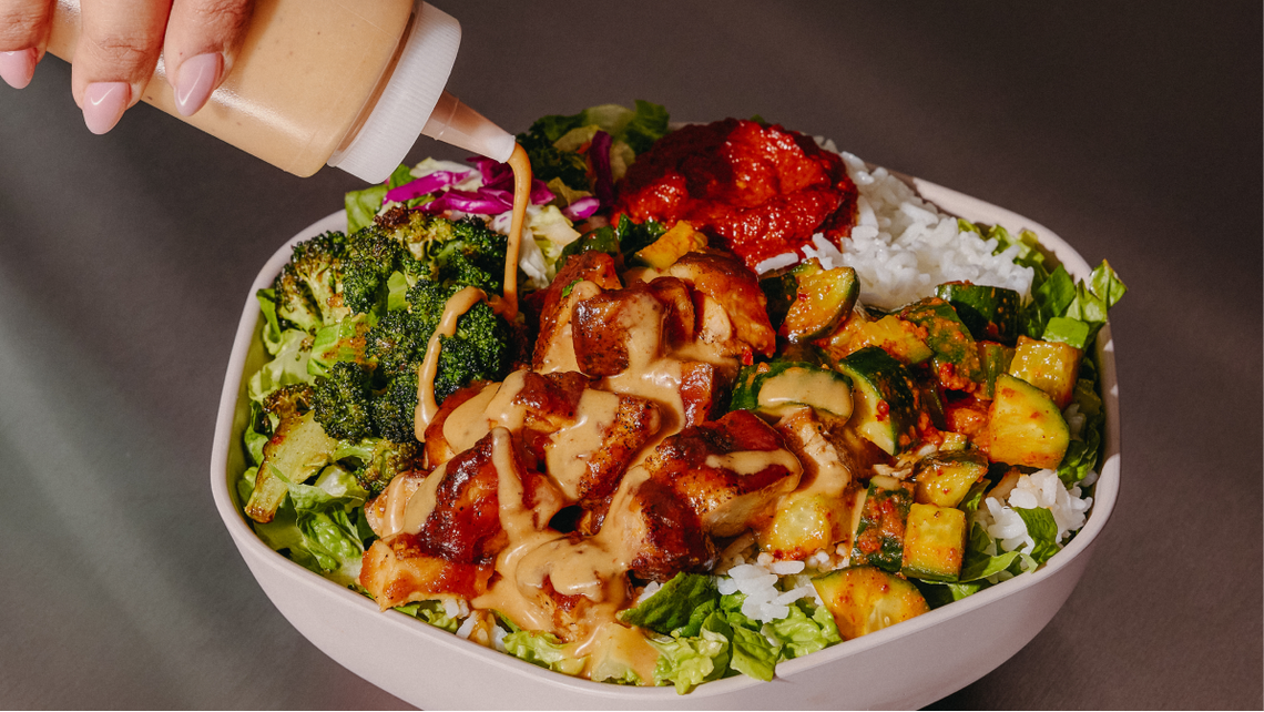 A hand squeezes a creamy orange dressing from a clear bottle onto a loaded grain bowl. The bowl is packed with white rice, grilled chicken, charred broccoli, purple cabbage, and a fresh cucumber salad with a side of spicy red harissa.