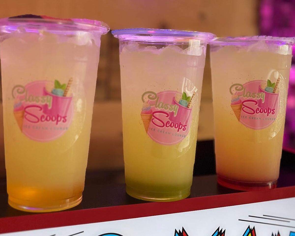 Three clear plastic cups containing a yellow and greenish layered beverage sit on a tray with a red border. Each cup has a “Classy Scoops Ice Cream Lounge” logo sticker on the side.