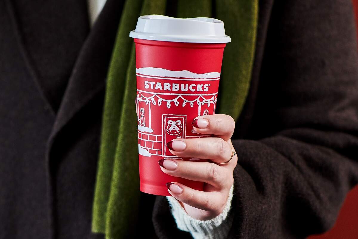 A person wearing a dark coat and a green scarf holds a reusable red Starbucks holiday cup featuring a festive white illustration of a building facade. The individual’s hand, with manicured nails, is visible holding the white lid of the cup, set against a red background.