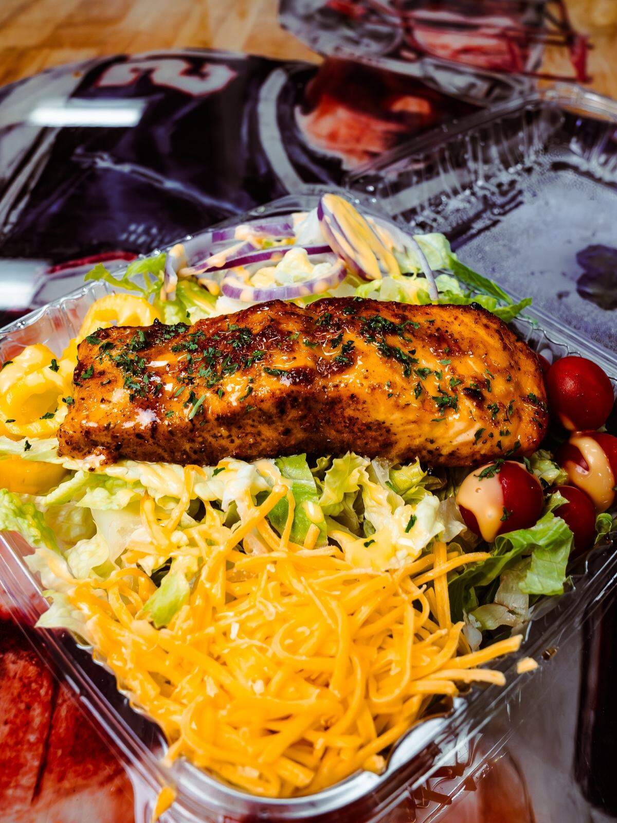 A seasoned, grilled salmon fillet rests on a bed of fresh greens, shredded cheddar cheese, and cherry tomatoes in a clear plastic container. The salad is lightly drizzled with a creamy dressing and garnished with fresh herbs.