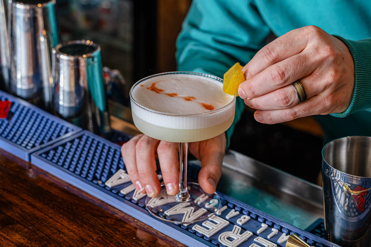 A bartender wearing a teal sweatshirt carefully garnishes a sophisticated cocktail at a wooden bar. The drink is served in a ribbed coupe glass, featuring a clear yellowish liquid topped with a thick, frothy white foam layer and a delicate drizzle of red bitters. The bartender’s hand, sporting a dark textured ring, gently pinches a vibrant yellow lemon twist onto the rim of the glass.