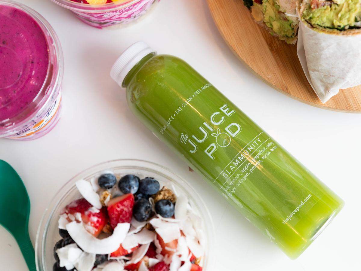 An overhead shot shows a bottle of “Glammunity” green juice centered among various healthy options, including a berry bowl and an avocado wrap. The flat-lay composition highlights the fresh, raw ingredients like kale, spinach, and coconut shavings on a clean white surface.