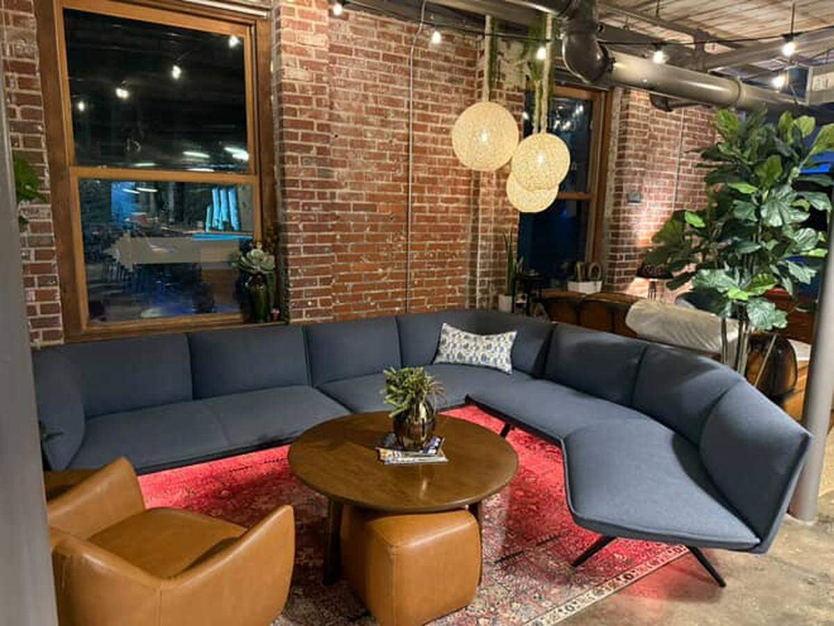 A comfortable lounge area features a large, curving, blue sectional sofa arranged around a circular wooden coffee table. The seating area is set on a patterned red rug and accented by hanging globe lights, exposed brick walls, and tall plants.