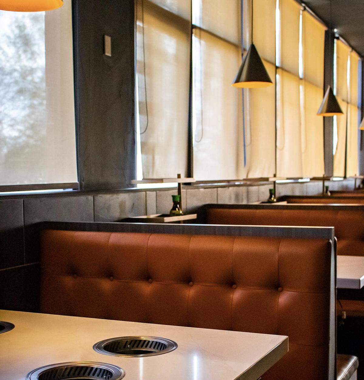 A restaurant interior with rows of deep reddish-brown leather upholstered booths and light marble-like tables with built-in circular grill vents. Cone-shaped pendant lights hang from the ceiling, and tall windows are covered by light beige roller blinds.