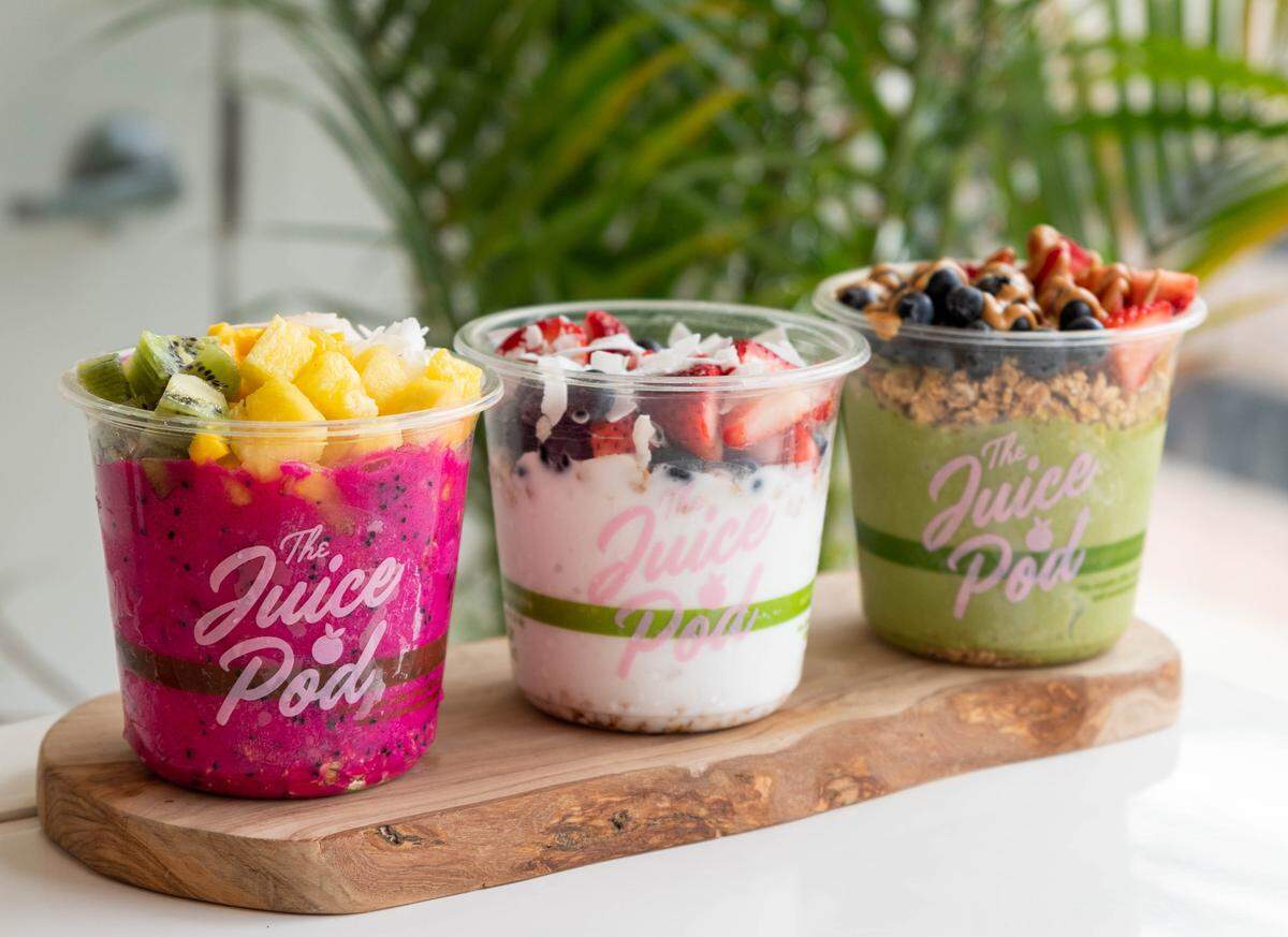 Three colorful smoothie bowls from The Juice Pod are lined up on a rustic wooden serving board. The bowls feature vibrant pink dragon fruit, white coconut and berry, and green superfood bases, each topped with fresh fruit, nuts, and granola.