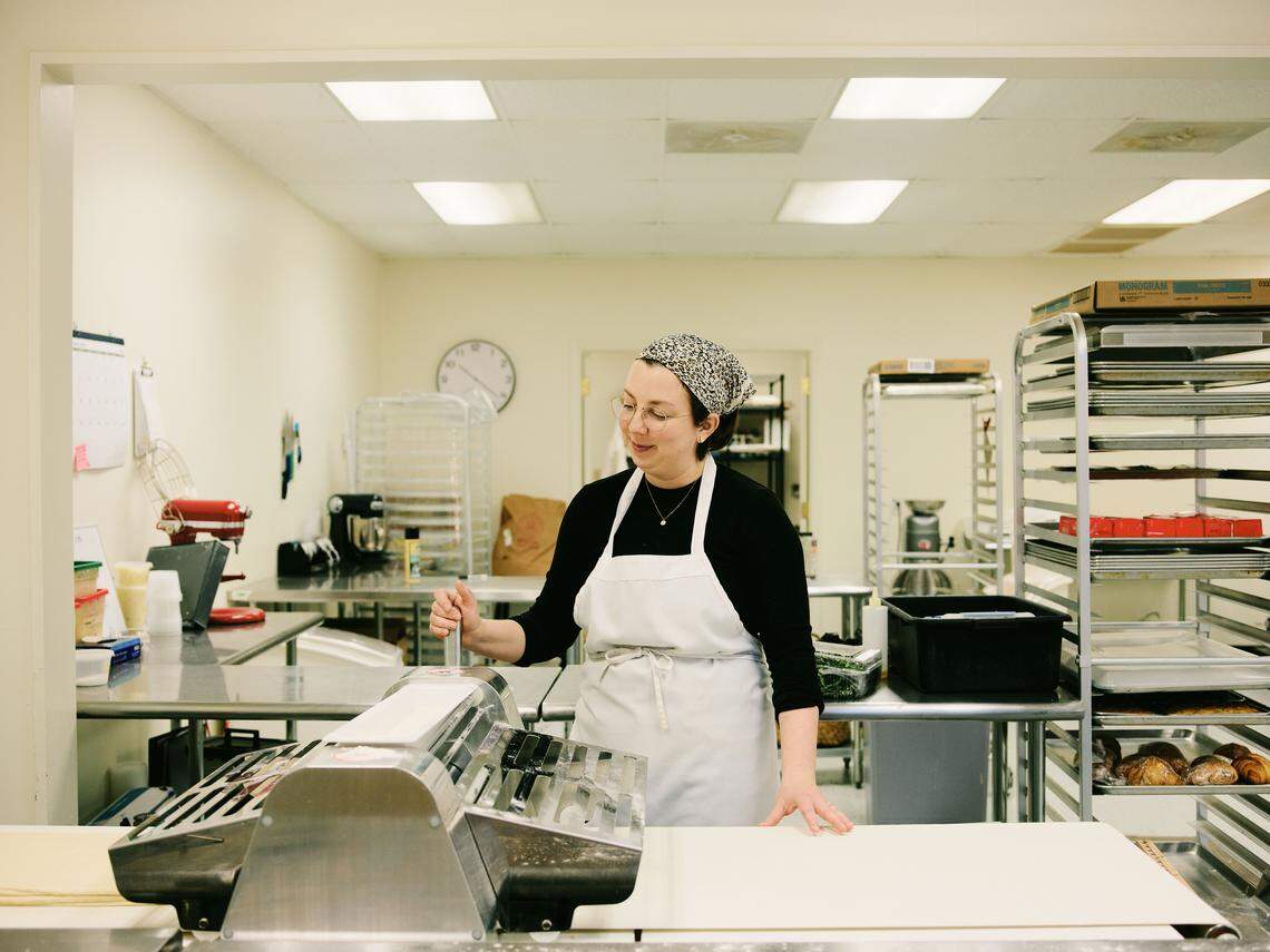 A woman wearing a white apron and a patterned headscarf stands in a professional bakery kitchen using a manual dough sheeter. She has a focused expression as she guides a long sheet of pale dough through the rollers on a stainless steel workstation. The background is filled with industrial kitchen equipment, including wire cooling racks, a red stand mixer, and various baking supplies.