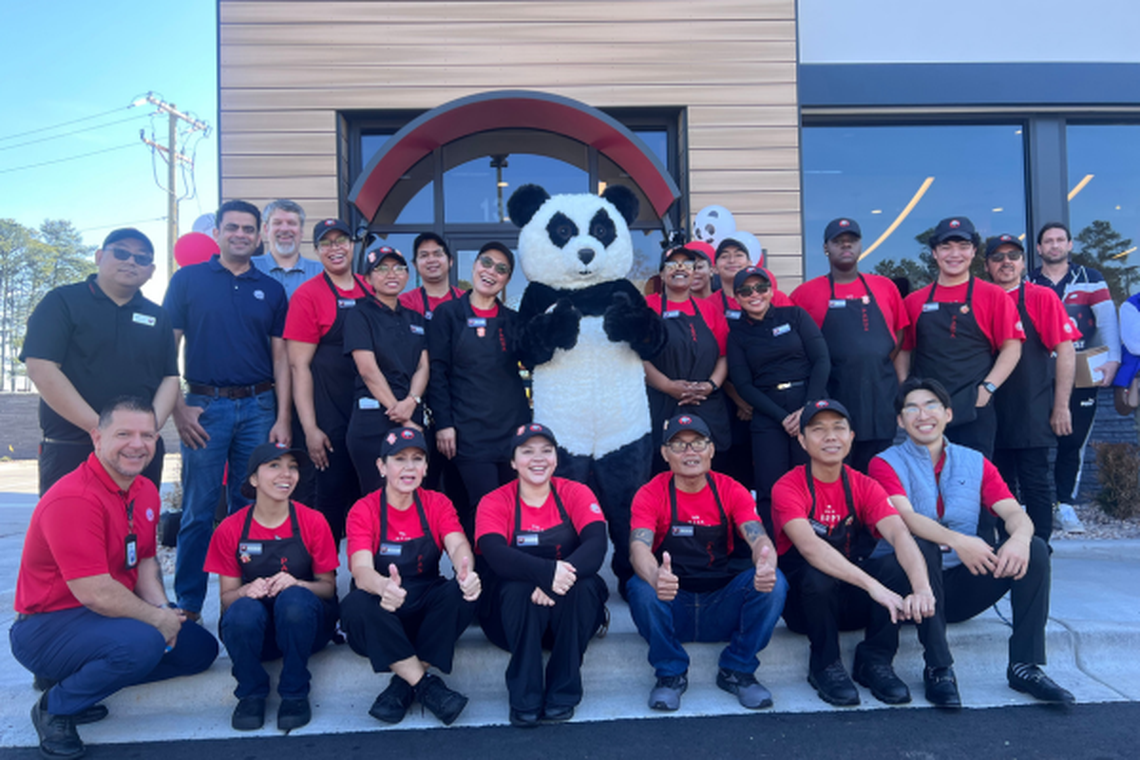 A group of about twenty Panda Express employees in red and black uniforms poses for a celebratory photo outside a restaurant alongside a large panda mascot. Several team members are sitting on a curb in the foreground giving thumbs-up gestures, while the rest of the staff stands behind them in front of the building’s entrance under a bright, clear sky.