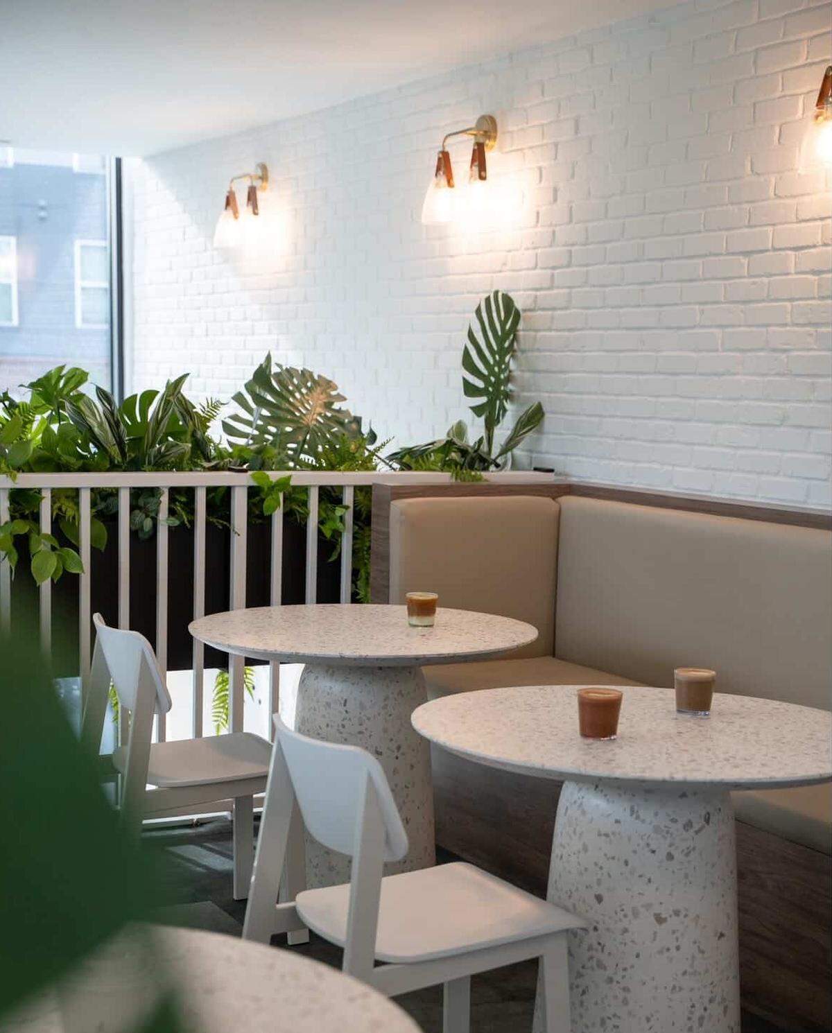 The interior of a bright cafe features white brick walls adorned with elegant gold wall sconces and lush tropical greenery. Two round, speckled terrazzo tables are paired with a tan built-in banquette and minimalist white chairs. Several iced coffee drinks are placed on the tables, creating an inviting and contemporary atmosphere.
