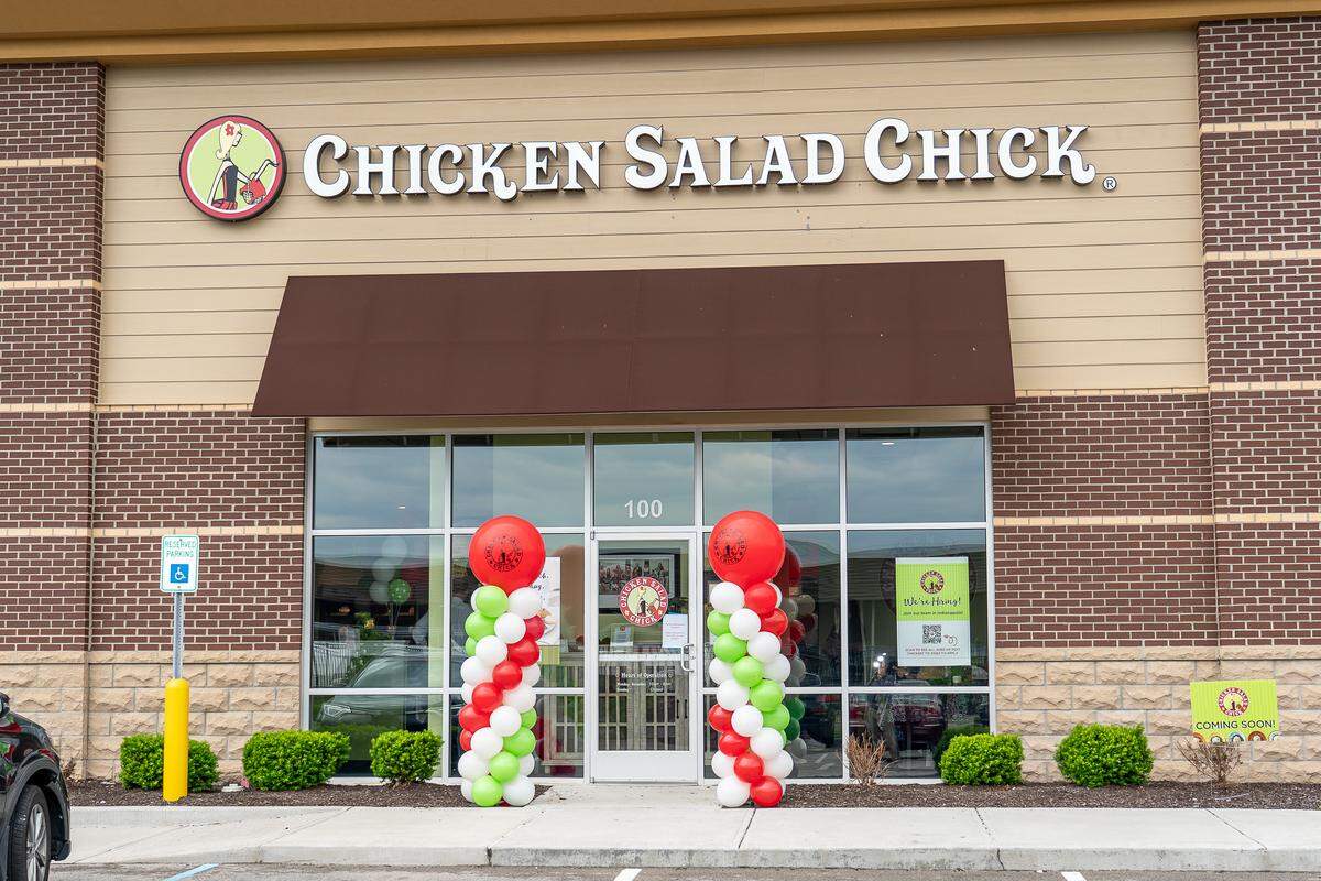 The exterior of a Chicken Salad Chick restaurant features a tan and brick storefront with a large white sign above the entrance. Festive red, white, and green balloon columns flank the glass front doors to celebrate a special event or opening.