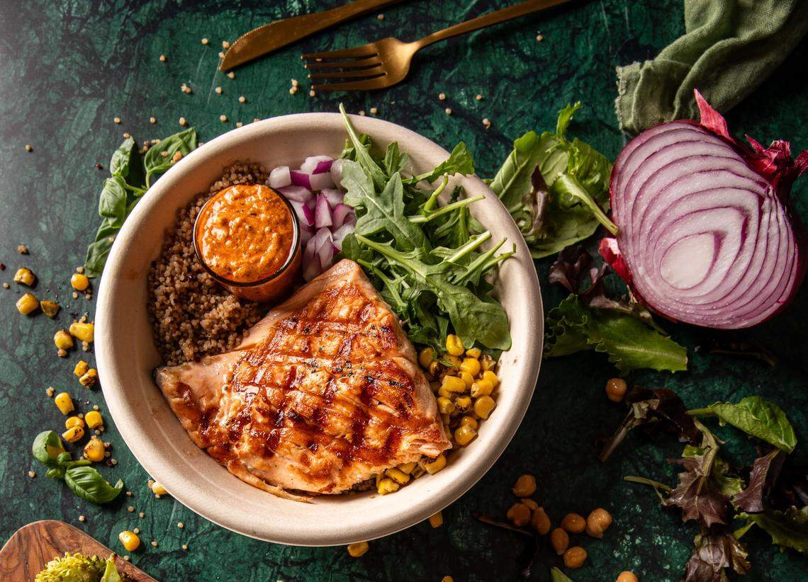 A top-down view of a healthy grain bowl containing grilled fish or chicken, quinoa or brown rice, corn, mixed greens, diced red onion, and a small cup of sauce. The bowl is set on a dark green surface next to gold silverware and a cross-section of red onion.