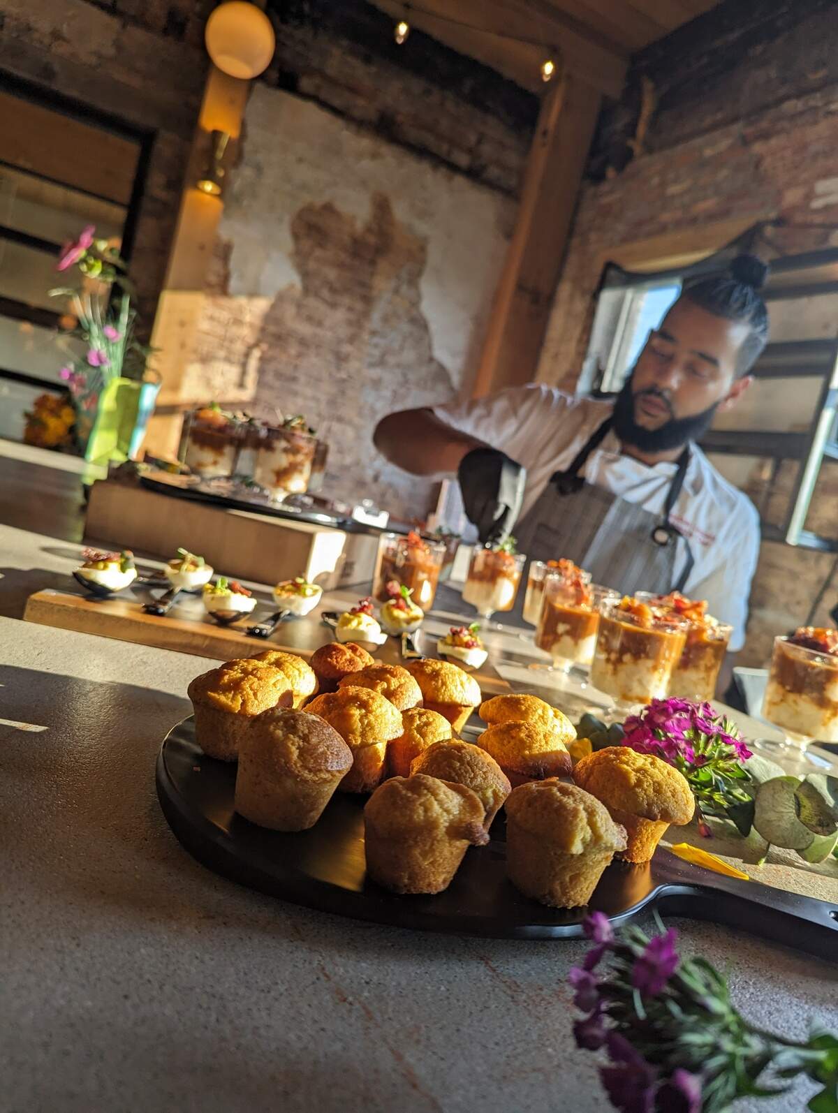A chef in an apron garnishes small dessert cups on a counter in a rustic, brick-walled room. In the foreground, a tray of mini muffins sits beside flowers in warm light.
