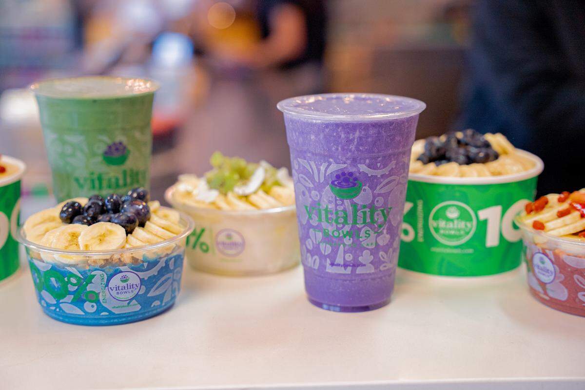 A row of colorful Vitality Bowls products includes a bright purple smoothie and a green smoothie in clear plastic cups. Beside them are various açaí bowls topped with fresh fruit like bananas, blueberries, and kiwi.