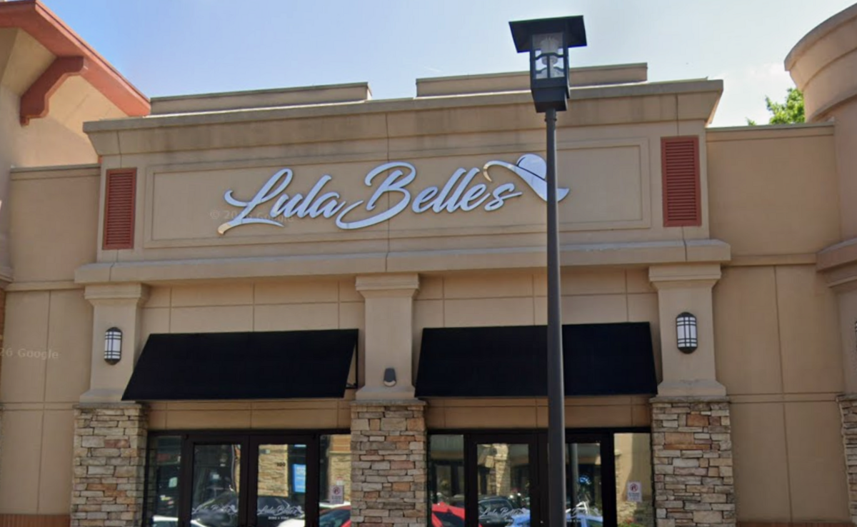 The exterior storefront of a boutique features a beige stone facade accented with stone-brick pillars and black fabric awnings over the windows and doors. Prominently displayed above the entrance is a large, white cursive sign that reads “Lula Belle’s,” with the apostrophe creatively styled as a wide-brimmed sun hat. A tall, black street lamp stands in the foreground on the right, partially obscuring the right side of the building’s signage.