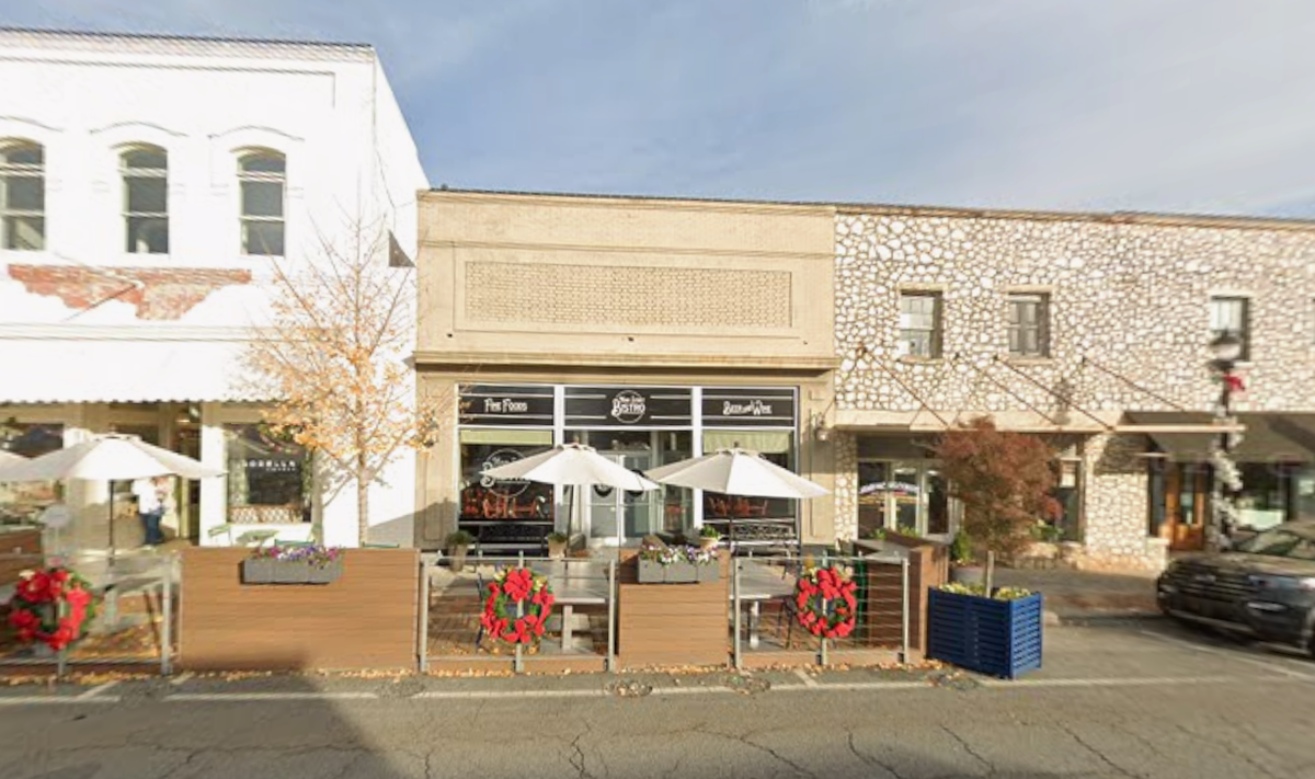 A storefront for “Main Street Bistro” features a modern black facade with white lettering and an outdoor dining area enclosed by wooden planters. Festive red wreaths and flower boxes decorate the patio, which is shaded by several white umbrellas. The restaurant is nestled between a historic white brick building and a textured stone building on a quiet city street.