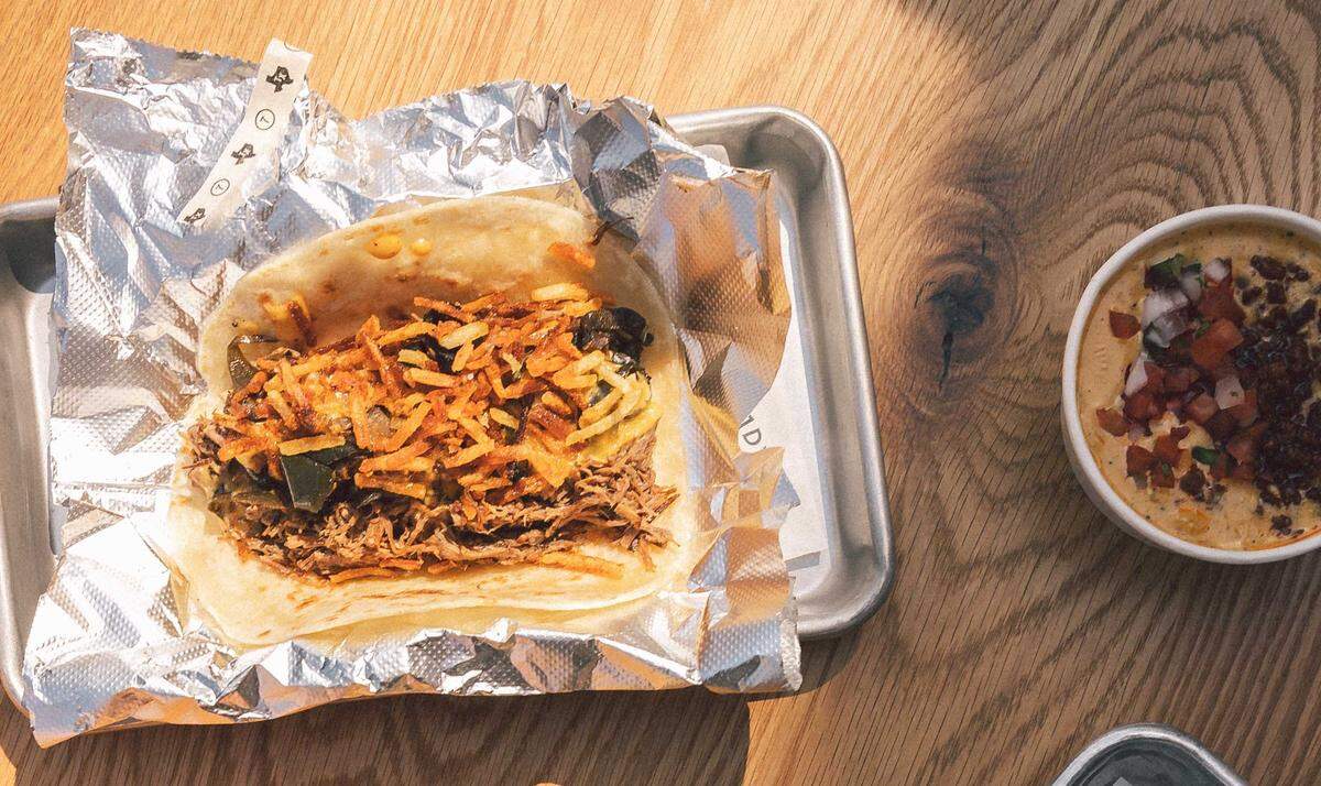 A close-up of a street taco on a metal tray, filled with shredded beef, dark green peppers, and a generous topping of crispy potato sticks. Beside the tray sits a small bowl of creamy white queso topped with fresh pico de gallo and crumbled chorizo.