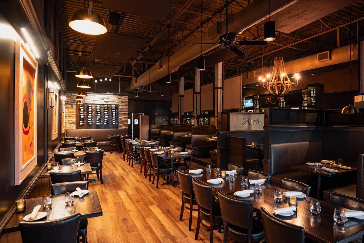 A wide view of a modern, upscale dining room featuring dark wood floors, black leather chairs, and neatly set tables with white linens. Industrial-style ceilings with exposed ductwork contrast with warm pendant lights and a large wooden chandelier. A stone accent wall in the back displays a large wine rack, while vibrant, abstract artwork in orange and yellow tones hangs on the dark left wall.