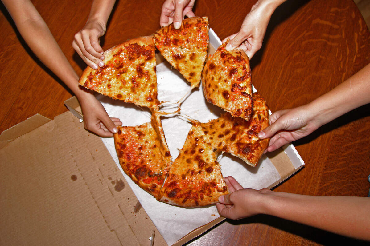 A high-angle shot captures multiple hands reaching into an open cardboard pizza box sitting on a wooden table. The hands are simultaneously pulling away six separate slices of a golden-brown cheese pizza, creating thin strands of melted cheese stretching toward the center. The composition focuses entirely on the shared meal, highlighting the casual, communal moment of enjoying a fresh pizza together.
