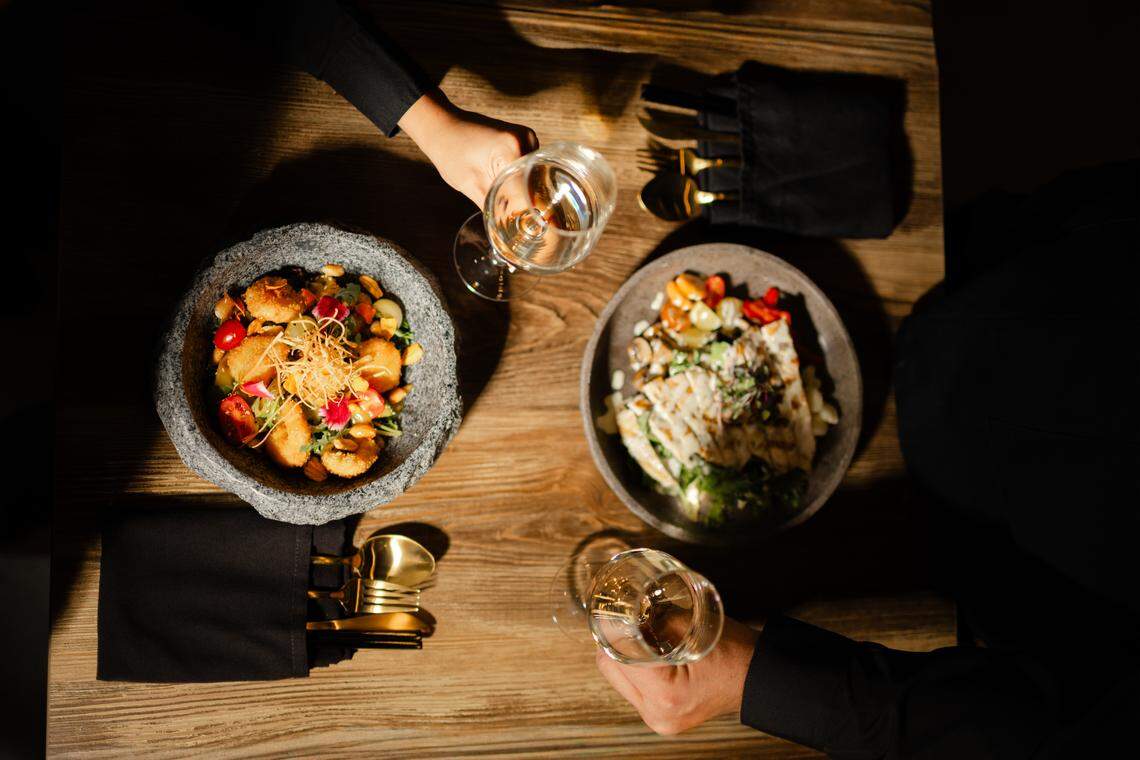 A dark overhead view of two people at a wooden table sharing two different salads and toasting with wine glasses. The left bowl contains a colorful “My Favorite Salad” with fried items and vegetables, while the right plate holds the “Lima Salad,” which appears to have grilled white fish or cheese on top. The photo captures a fine-dining ambiance with golden utensils visible in a dark napkin holder.