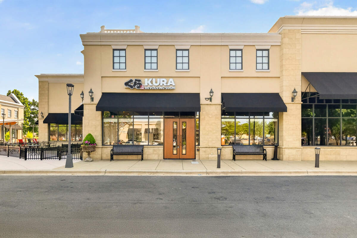 The exterior storefront of a Kura Revolving Sushi restaurant is housed in a modern beige stucco building with black fabric awnings over large glass windows. Double wooden doors mark the central entrance, flanked by black benches and classic lamp posts on a clean concrete sidewalk. In the background to the left, an outdoor patio dining area with black fencing and red chairs is visible under a clear blue sky.